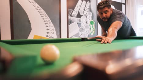 young bearded man sinking red ball number three into a pocket casual sports game of billiards on a green cloth