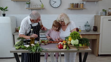 elderly grandparents in kitchen teaching grandchild girl how to cook salad, chopping red pepper