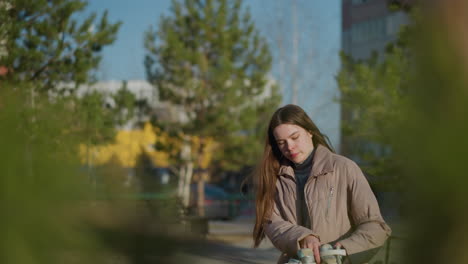 a young woman in a peach jacket and grey inner shirt walks through a park on a sunny day, holding something in her hand, as she sits on a bench. the background includes greenery and buildings