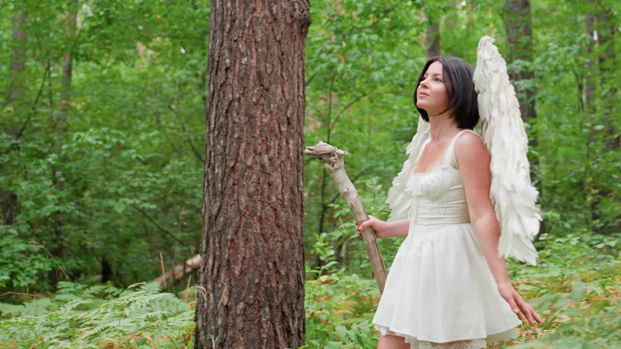 Female goddess with white wings walks through lush woodland holding wooden staff, surrounded by green ferns and towering trees, radiating grace, strength, purity, and mystical presence