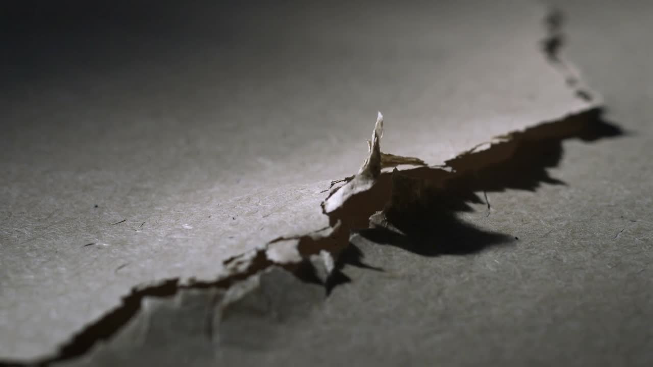 Widening jagged tear on cardboard causing torn flap to lift and cast deep shadow on tabletop