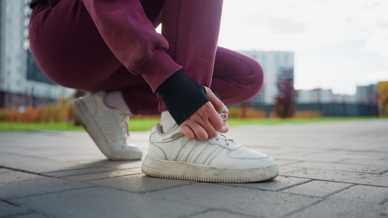 Woman bends down to tie canvas sneaker lace on paved urban sidewalk outside modern apartments under warm calm morning light showcasing focused preparation for outdoor fitness routine