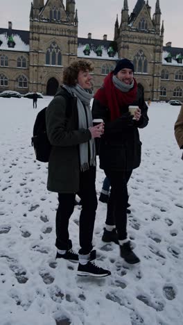 students walking together across snow covered university grounds, sipping hot beverages from disposable cups, sharing conversation with historic academic building framing winter landscape backdrop