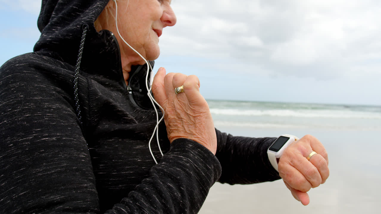 Side view of old caucasian senior woman using smartwatch and listening music on earphones at beach 4