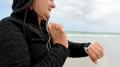 Seitenansicht-Einer-Alten-Kaukasischen-Seniorin,-Die-Am-Strand-4-Eine-Smartwatch-Benutzt-Und-über-Kopfhörer-Musik-Hört