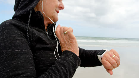 vista lateral de una anciana mujer caucásica usando un reloj inteligente y escuchando música en auriculares en la playa 4