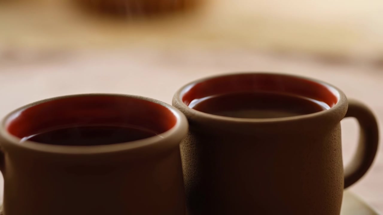 Dos tazas de cerámica marrones con asas reposan una al lado de la otra sobre una superficie de mesa clara en primer plano, con un fondo interior suavemente desenfocado y una cálida iluminación natural.