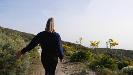 mujer joven camina por un camino rebosante de vegetación a los lados y toca arbustos con las manos