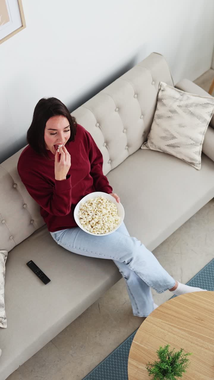 Ung kvinde slapper af på sofaen med popcorn og fjernbetjening