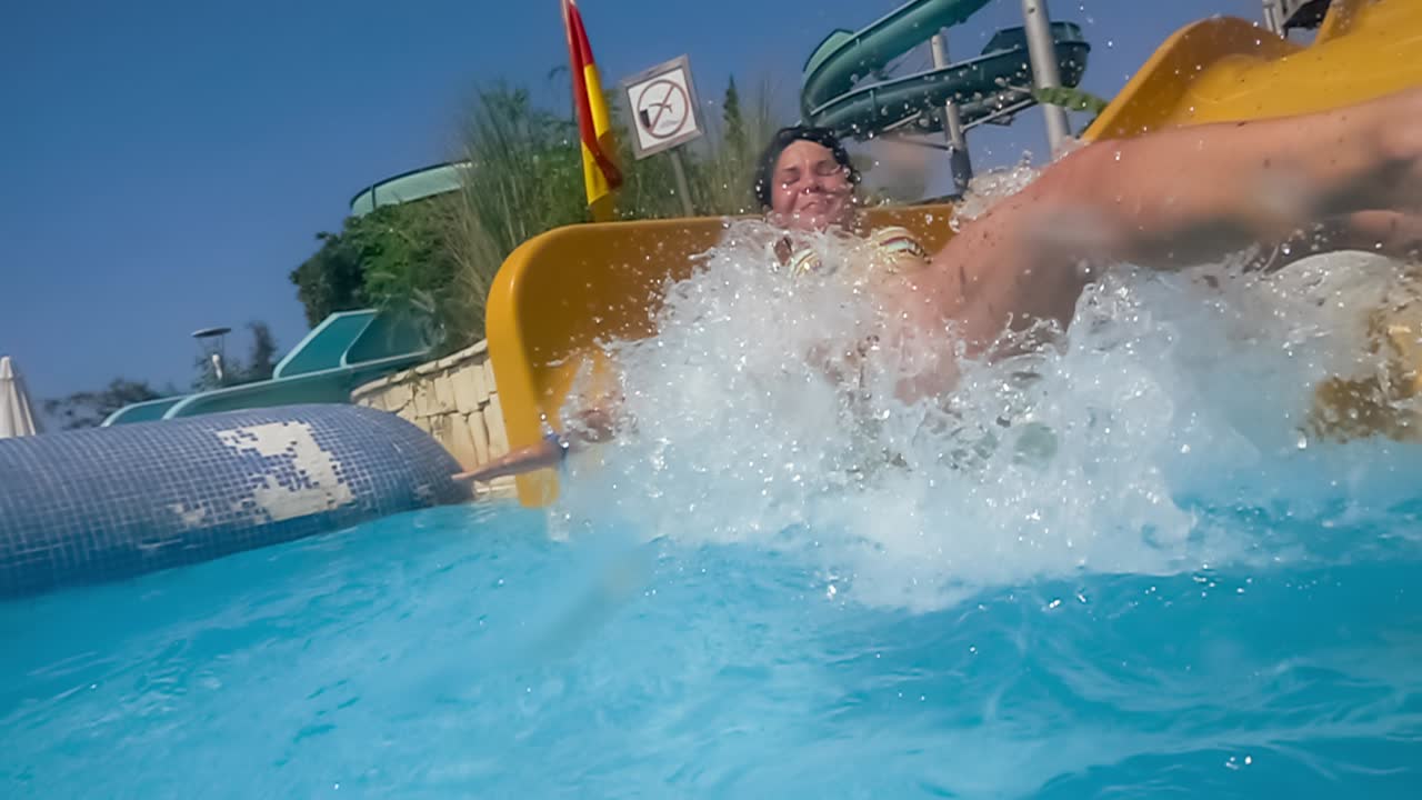 descenso del tobogán acuático en un parque acuático de vacaciones. cámara lenta en un tobogán acuático vacaciones familiares, una mujer en bikini desciende del tobogán en una piscina de agua azul salpicando gotas de agua.