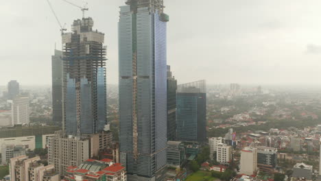 Toma-Aérea-De-Carro-Volando-Hacia-Dos-Rascacielos-De-Vidrio-En-Construcción-En-Un-Denso-Centro-Urbano-En-Yakarta