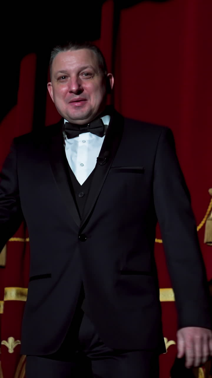 Portrait of a host. Man actor in black costume appears on stage and begins the speech in bright light spot in the theater. Vertical video