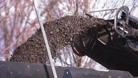 slow motion footage of ground up road asphalt being churned out of a machine and into the back of a construction truck as traffic drives in the opposite lane