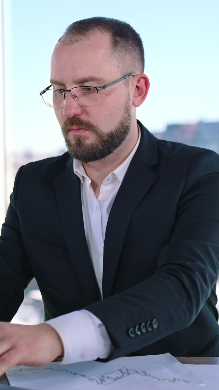 Serious business worker on office window backdrop. Middle aged businessman in glasses sitting at desk in his workplace. Entrepreneur searches solution of business issues. Vertical video
