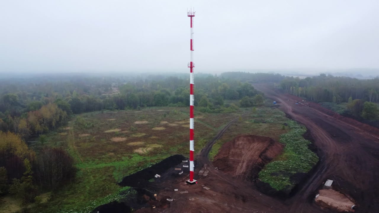 Pohled z dronu ukazuje aktivní staveniště na otevřeném poli vedle zeleně, s hliněnou zemí, dřevěnými prkny, hromadami zeminy, roztroušeným materiálem a tlumeným zataženým světlem, s důrazem na stavbu, letecký pohled a samotné staveniště.