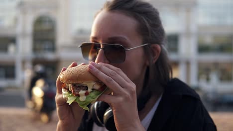 mujer joven atractiva, sonriendo alegremente, sostiene una sabrosa hamburguesa en dos manos y la come. vestida con ropa casual, con gafas de sol