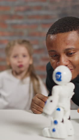 Cheerful guy and Asian friend look at cute dancing robot at table while girl student jumps copying toy moves in studio closeup