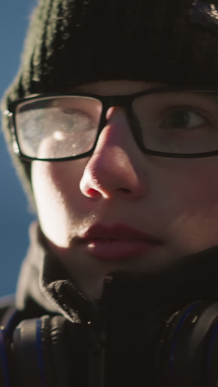 Close-up of a young boy wearing glasses and a black jacket adjusts his glasses, looking thoughtful, he has a headset around his neck