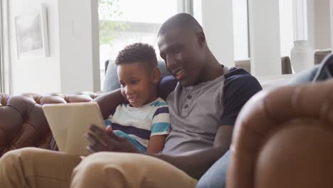 video of happy african american father and son sitting on sofa and using tablet