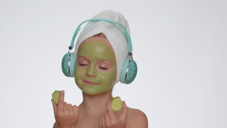niña sonriente hidratante máscara de pepino verde en la cara escuchando música en los auriculares bailando