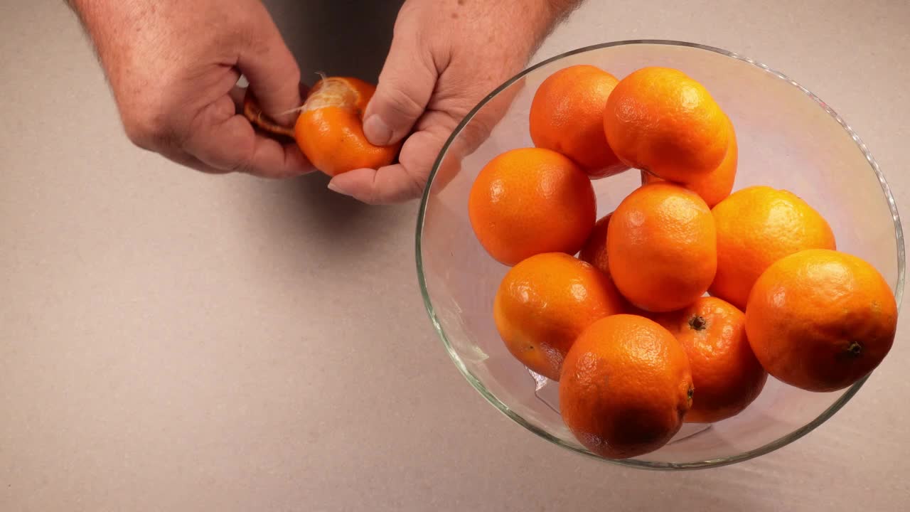 las manos de un hombre mayor recogiendo un satsuma de un cuenco y luego pelándolo y poniéndolo entero.