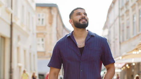 Happy-young-adult-indian-man-tourist-walking-in-urban-city-street,-smiling-having-positive-good-mood