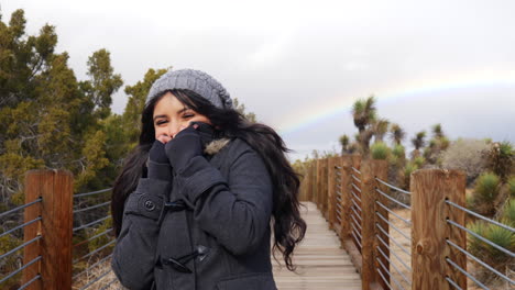 attractive young woman smiling in a cold winter rain storm with a colorful rainbow in the cloudy sky slow motion