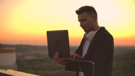 a man in headphones on the roof relaxes working remotely enjoying life despite a handsome kind of sipping beer and types on the keyboard. trade on the stock exchange using a laptop and enjoying the beautiful view