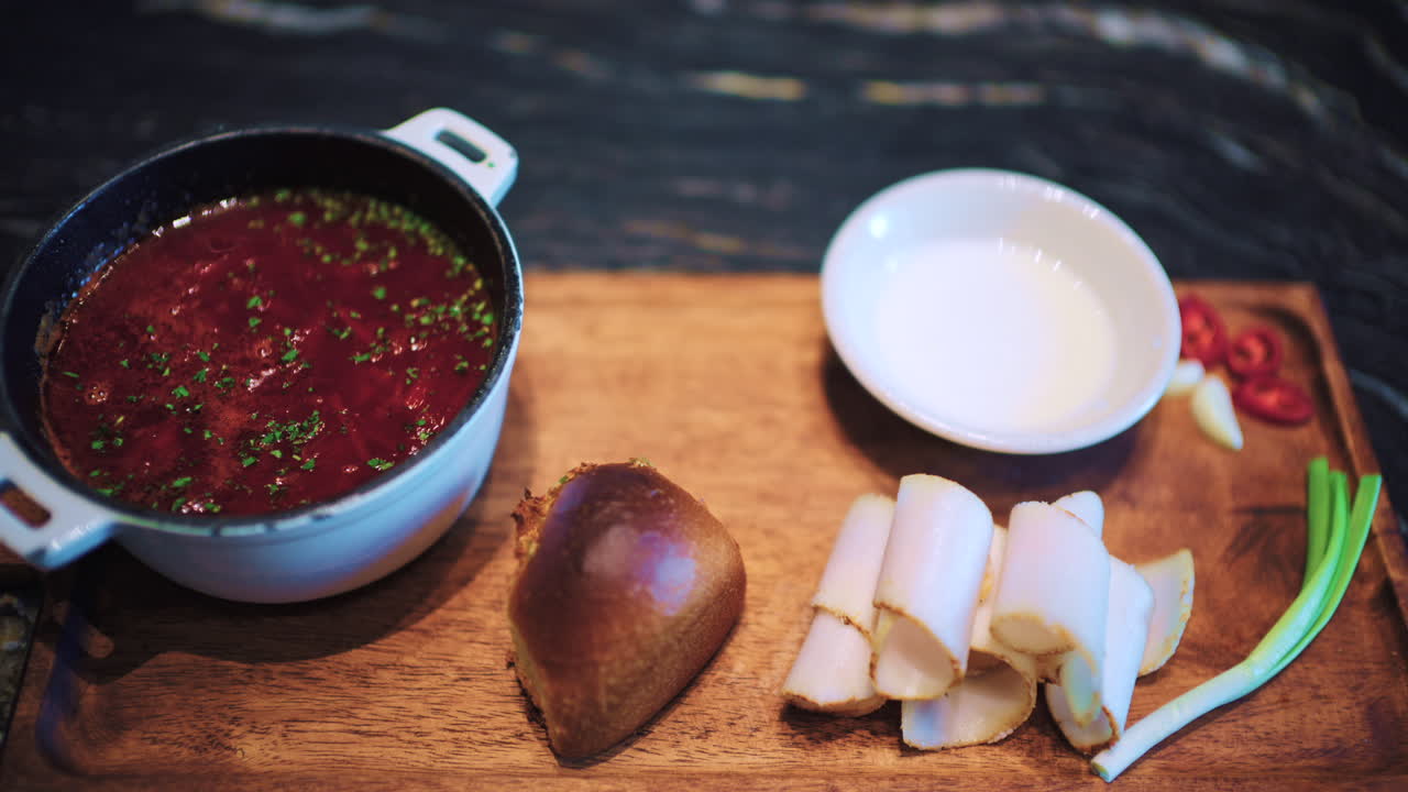 Nahaufnahme einer Schüssel mit Borschtsch, serviert auf einem Holzbrett zusammen mit Brot, geschnittenem, geräuchertem Speck und Frühlingszwiebeln.