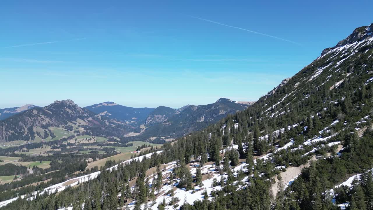 Drone view of Oberjoch in the Bavarian Alps showing snow patches, green meadows, and forested hills. A peaceful mountain village perfect for nature and travel visuals.