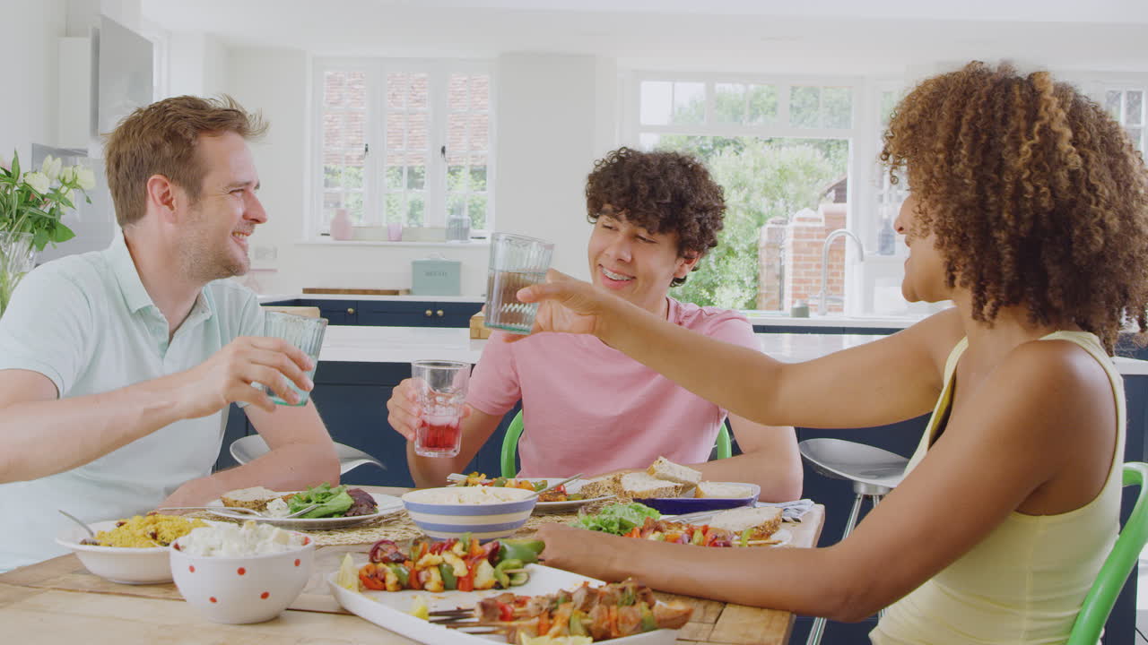 Multi-Racial Family With Teenage Son Sitting Around Table In Kitchen At Home Eating Meal Together