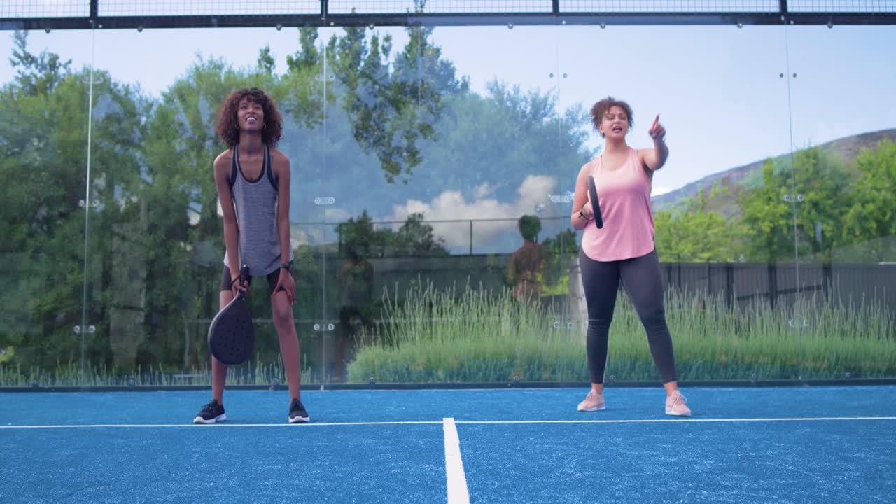 Afroamerikanska kvinnor värmer upp på en blå bana med racketar, där den högra kvinnan pekar, hoppar och svingar.