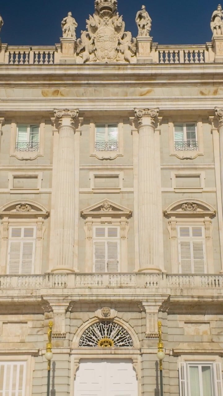 Ripresa verticale dal basso che mostra l'ornata facciata neoclassica del Palazzo Reale di Madrid. Cattura le dettagliate sculture in pietra, le statue e le imponenti colonne sotto un cielo azzurro limpido