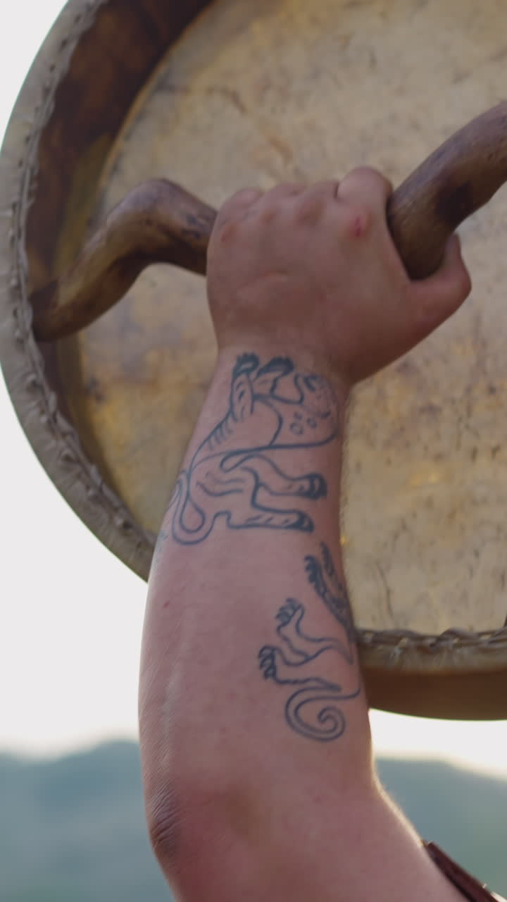Fat man with tattoos on arm raises up shamanic drum against mountains closeup. Altai religious rituals for spirits blessing. Meditative music