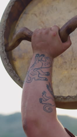 fat man with tattoos on arm raises up shamanic drum against mountains closeup. altai religious rituals for spirits blessing. meditative music