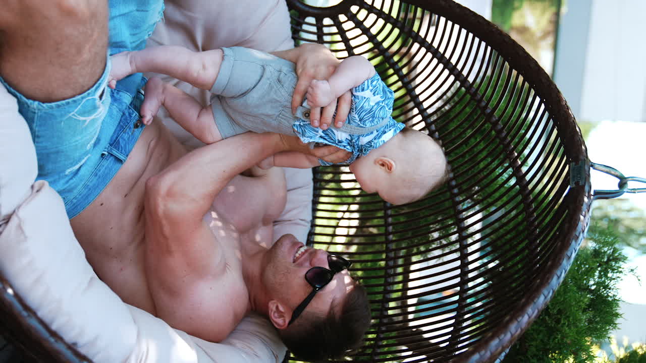 Caucasian man in jeans shorts sits in egg chair holding up his baby. Father talks to his infant son with a smile. Vertical screen.