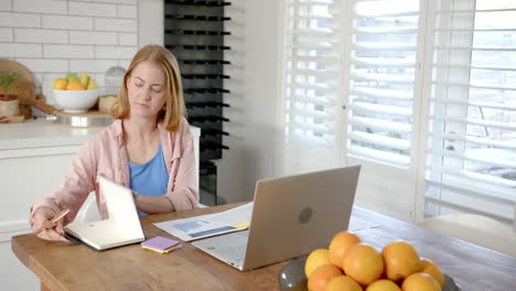 working from home, woman using laptop and writing in notepad at kitchen table
