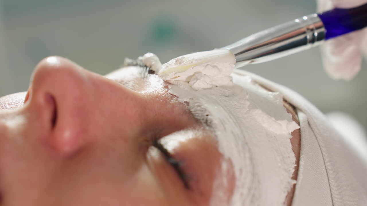 Hands of aesthetician in protective gloves spreading creamy mask in circular motion with silicone brush over forehead of resting model conveying gentle spa facial treatment under soft lighting