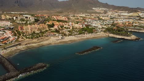 Erstaunliche-Drohnenaufnahme-Der-Küste-Am-Meer-Der-Stadt-Mit-Gebäuden-Mit-Bergen-Im-Hintergrund-Blaues-Wasser-Meer-Drohnenaufnahme-Palme-Panoramablick-In-Spanien-Teneriffa-Los-Cristianos