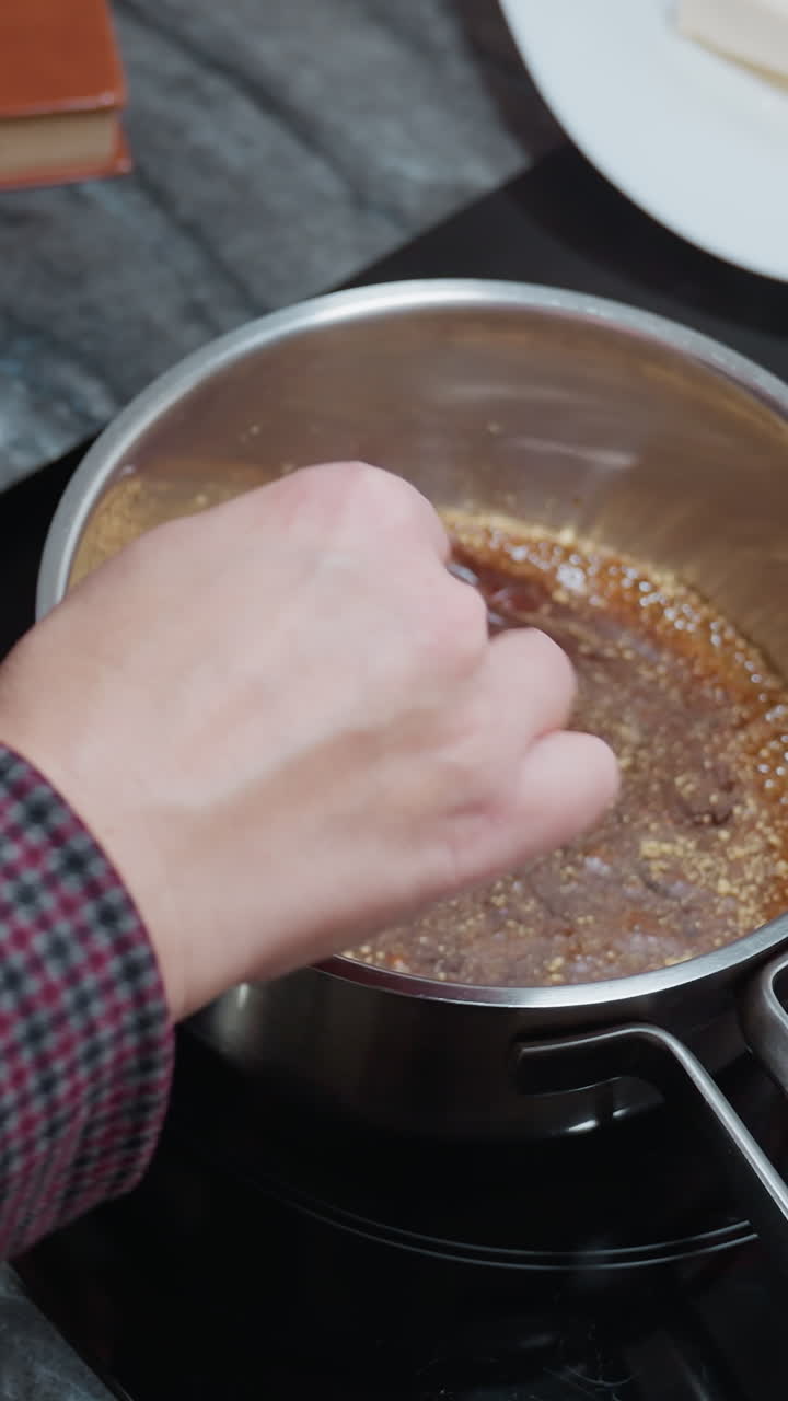 vista parcial de arriba hacia abajo de una persona agitando sopa en una olla de acero inoxidable usando un palo en una estufa eléctrica, con bandeja de madera, utensilios e ingredientes dispuestos en una encimera de mármol oscuro en una cocina moderna