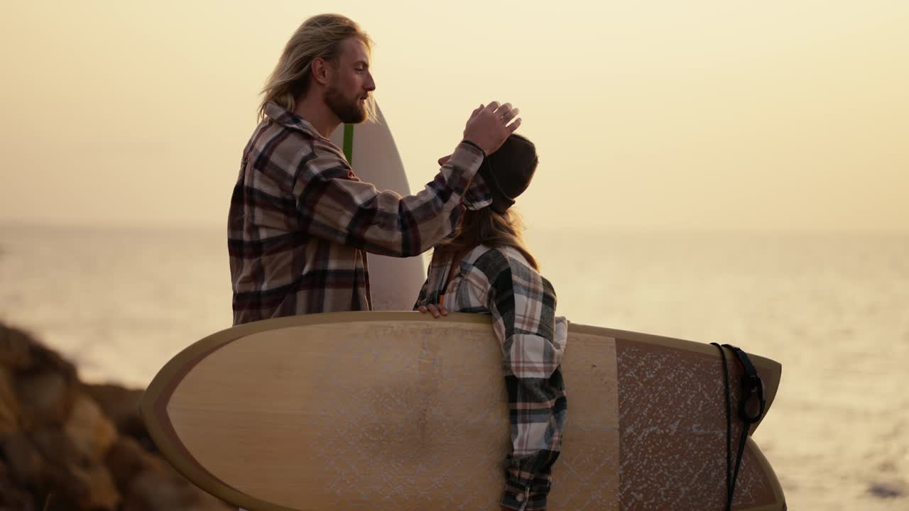 A blond guy with a beard in a plaid shirt straightens his hair and strokes the head of his blonde girlfriend in a black hat and plaid shirt who is holding a surfboard standing on a rocky shore near the sea in the morning