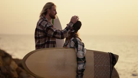 A-blond-guy-with-a-beard-in-a-plaid-shirt-straightens-his-hair-and-strokes-the-head-of-his-blonde-girlfriend-in-a-black-hat-and-plaid-shirt-who-is-holding-a-surfboard-standing-on-a-rocky-shore-near-the-sea-in-the-morning