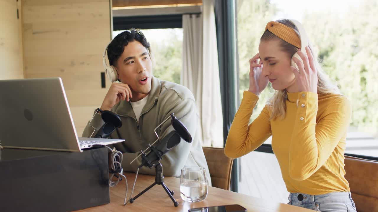 ポッドキャストスタジオで、アジア人男性と白人女性の共同ホストがヘッドホンを手に取りマイクのテストを行い、その後録音している。