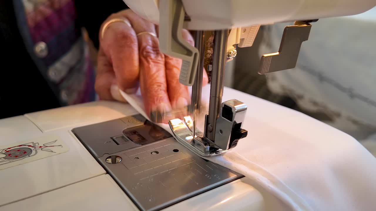 4K static footage of a woman’s hand guiding white fabric as it is stitched by an electric sewing machine. Ideal for craft, textile, fashion, production, and handmade industry projects