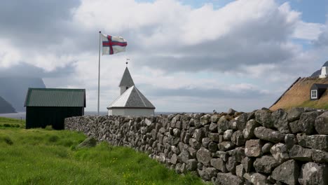 wide shot showing waving faroese flag in front of small church on faroe islands during cloudy day