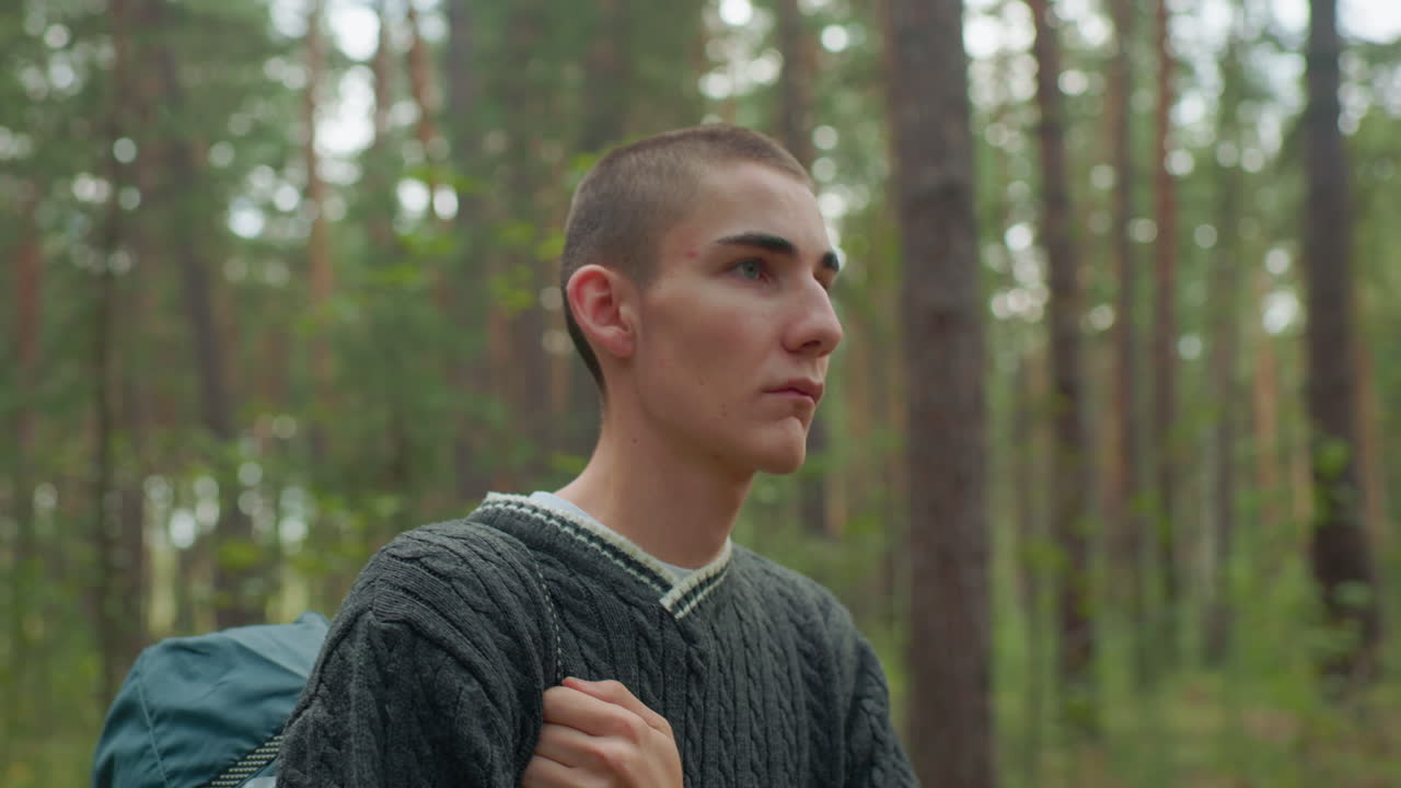 Young boy walking through forest path while holding strap of sleeping bag over shoulder, dressed in cozy sweater, appearing focused and calm as he travels alongside companion