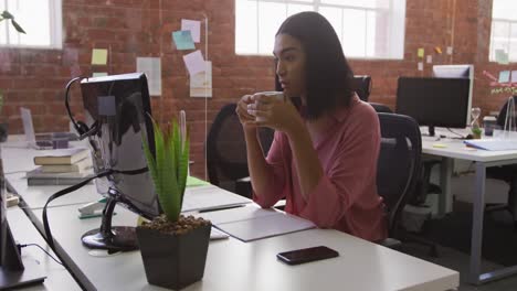 Mixed-race-businesswoman-sitting-at-desk-drinking-coffee