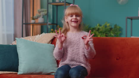 Portrait-of-little-child-girl-kid-alone-on-sofa-at-home-showing-ok-gesture,-like-sign,-positive-good