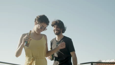 Cheerful-couple-holding-beer-and-dancing-at-rooftop-party