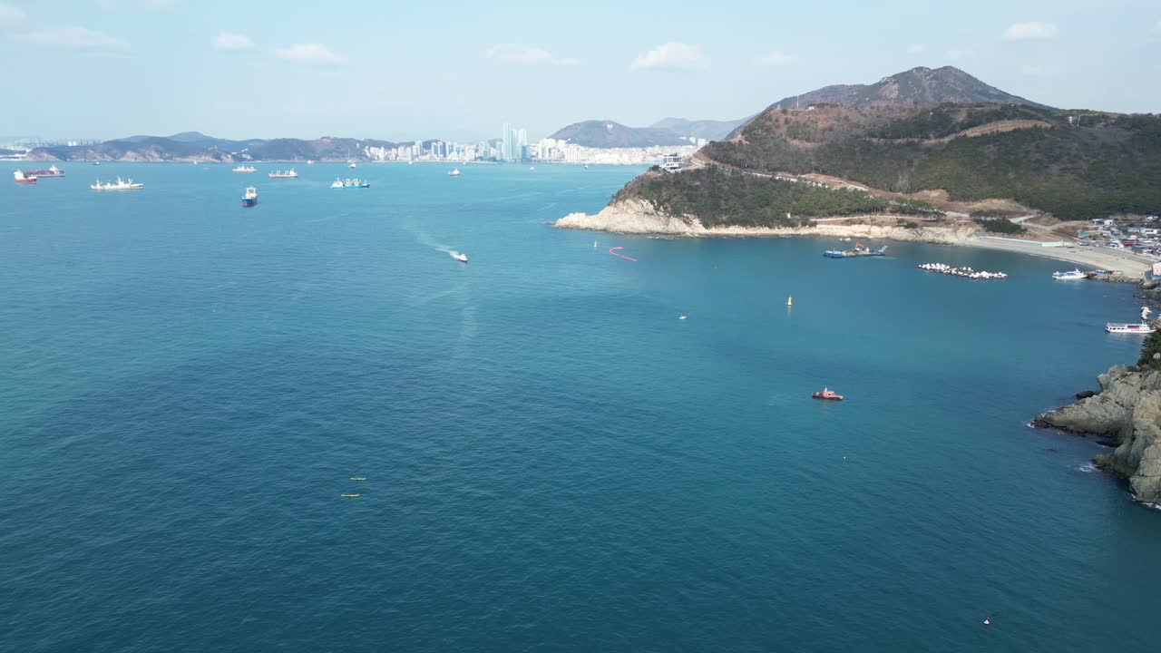 Drone aerial view in South Korea countryside flying over the dark blue sea of Busan next to a green mountain, ships and city in the background sunny day